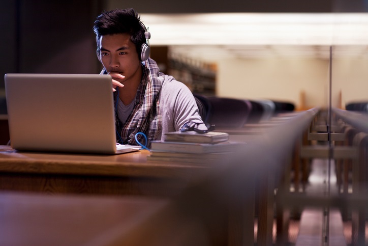 A youth is sitting at a desk in a library, looking at a laptop computer screen.